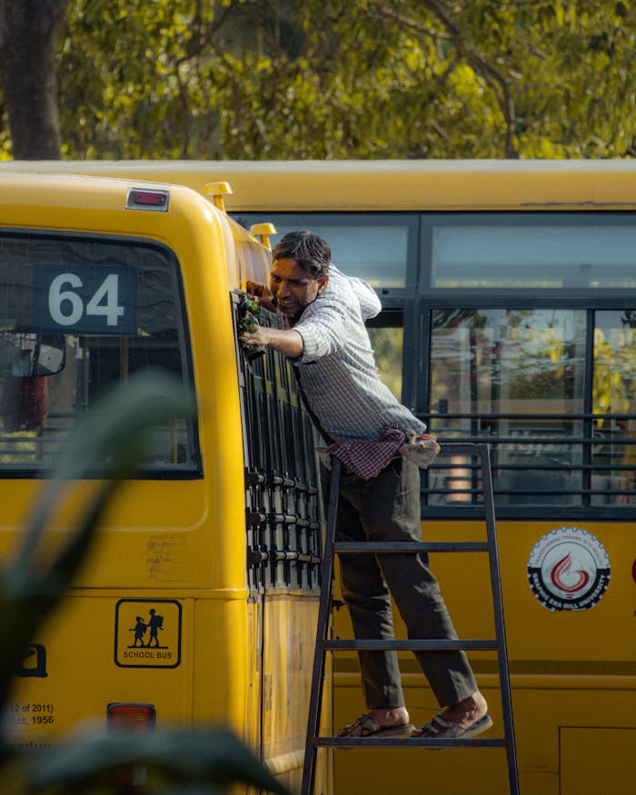 A worker on a ladder performing maintenance on a yellow school bus during the day.